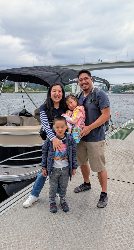 A smiling family of four stands on a dock beside a pontoon boat, ready for their Porto river cruise, with a bridge and river in the background.