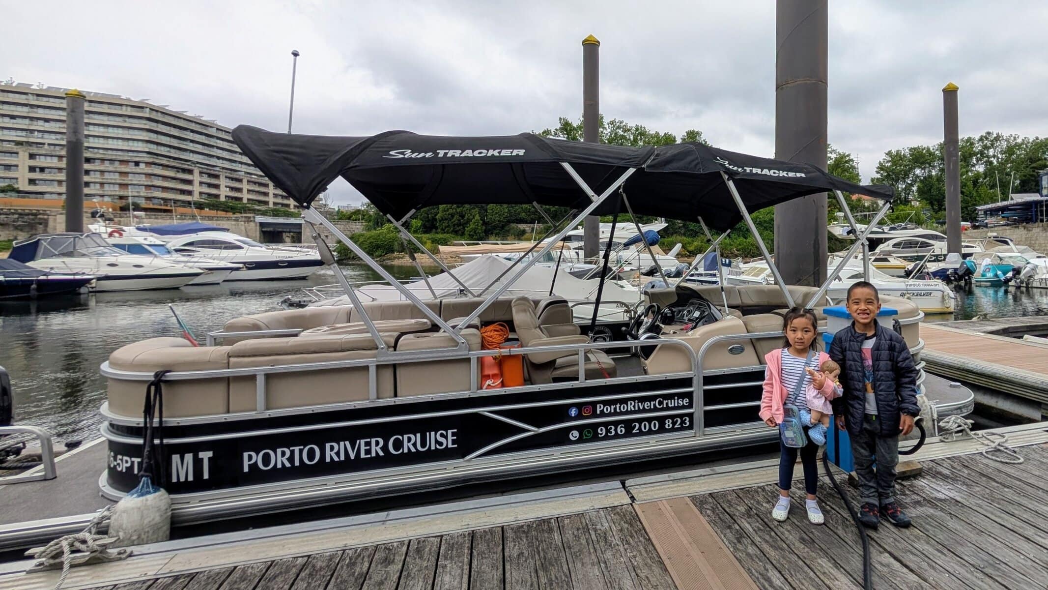 Two children stand on a dock beside a Porto river cruise boat, with several other boats and buildings visible in the background.