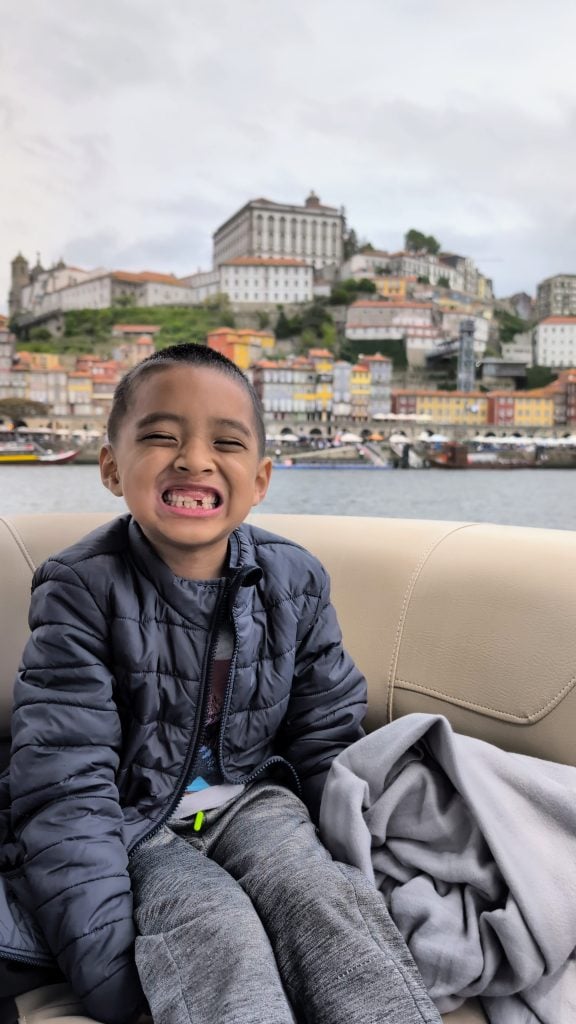 A young boy in a dark jacket smiles widely while sitting on a boat in Porto, with colorful buildings and a vibrant hillside cityscape in the background.