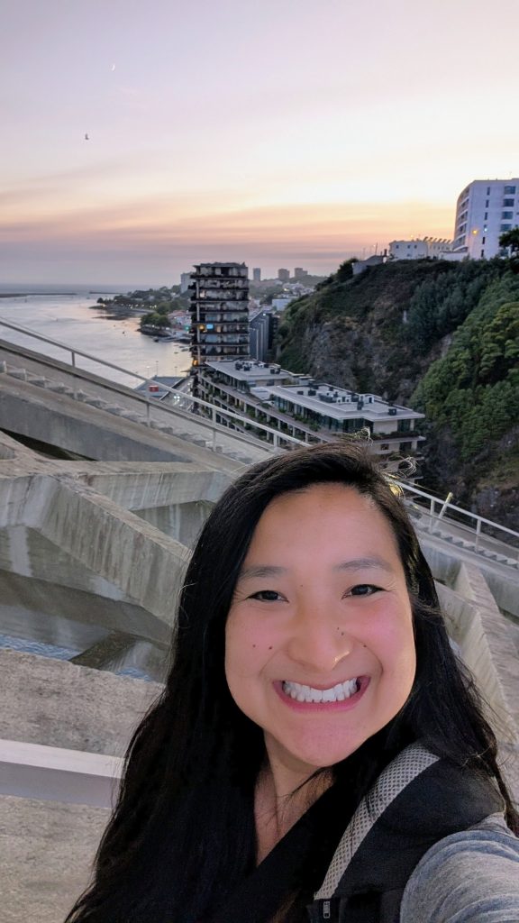A woman smiles for a selfie on a bridge at sunset, with buildings, trees, and a body of water visible in the background.