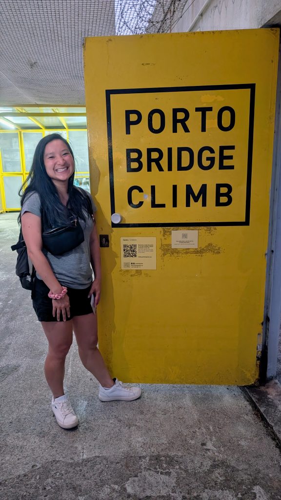 A woman stands smiling next to a bright yellow door labeled "Porto Bridge Climb" inside an indoor area.