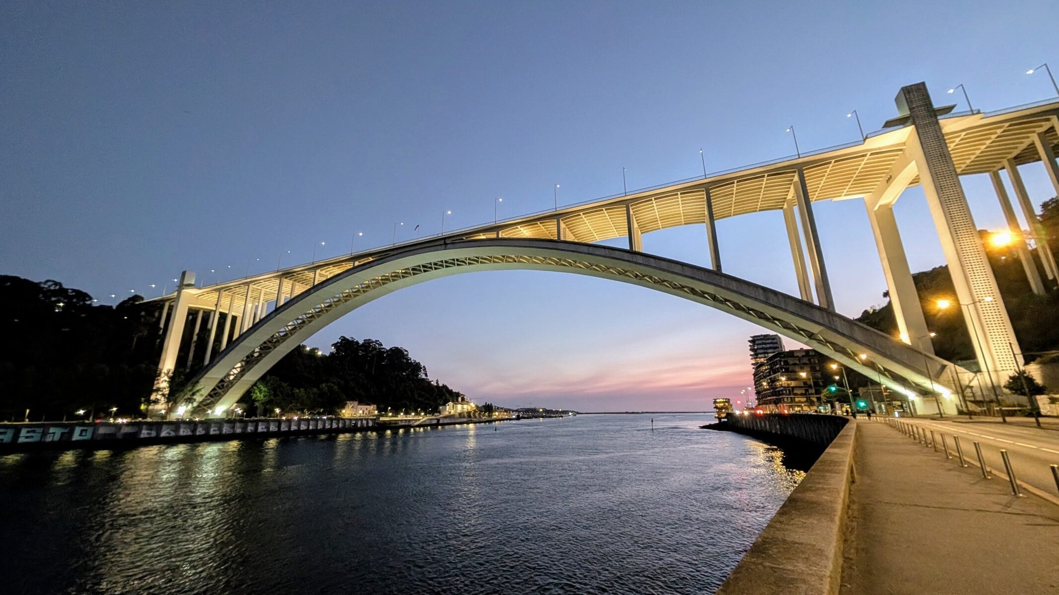 A large arched bridge, the Arrábida Bridge, spans a wide river at dusk, with streetlights on and buildings along the waterfront beneath a clear evening sky—an ideal scene for a Porto bridge climb.