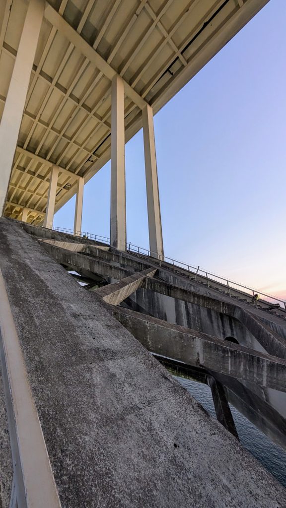 View from beneath the Arrábida Bridge, showcasing its massive support beams and tall vertical columns above the water below, all under a clear sky.