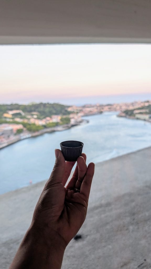 A hand holds a small chocolate cup filled with port wine with a blurred Douro River and the Porto cityscape visible in the background.
