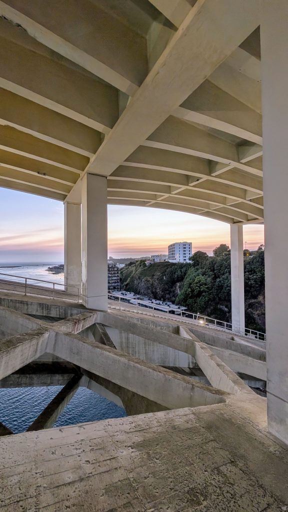 View of the ocean, coastline, and buildings through the structural beams of the Arrábida Bridge at sunset—a stunning perspective you can experience on a Porto bridge climb.
