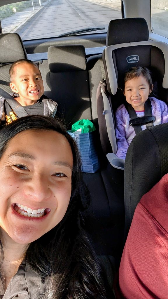 A woman takes a selfie in a car with two smiling children in the back seat, one in a car seat.