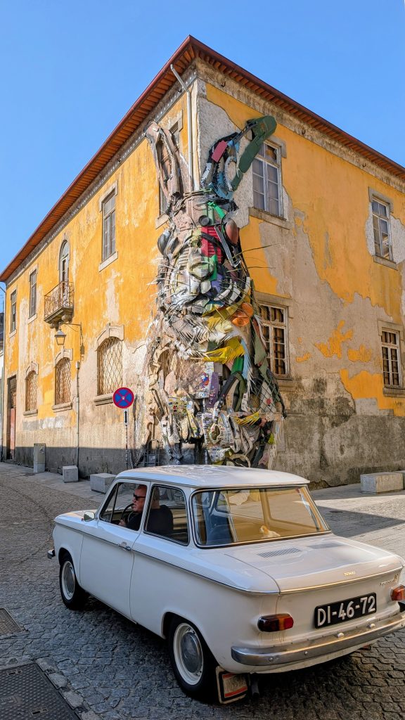 A white vintage car is parked on a cobblestone street in front of a yellow building with a large, colorful rabbit mural created from recycled materials, called Half Rabbit, in Gaia.