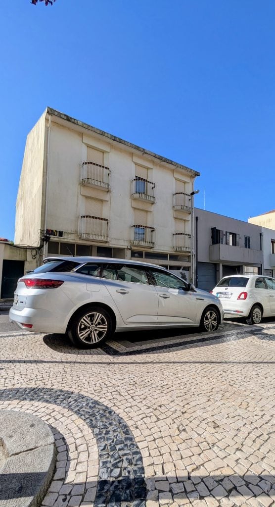 A white station wagon and a white compact car are parked on a cobblestone street in front of a three-story beige building with small balconies.