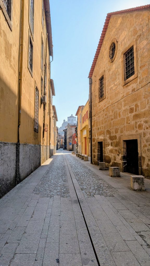 A narrow, empty cobblestone street in Vila Nova de Gaia lined with old stone and stucco buildings on a sunny day.