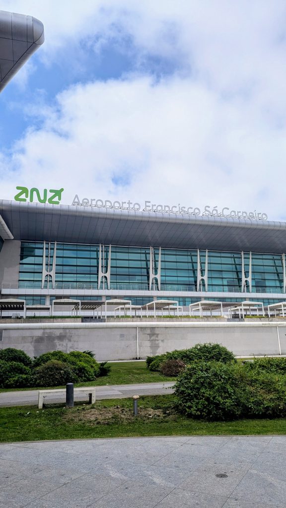 Exterior view of the ANA Aeroporto Francisco Sá Carneiro building with glass windows and green landscaping under a cloudy sky.