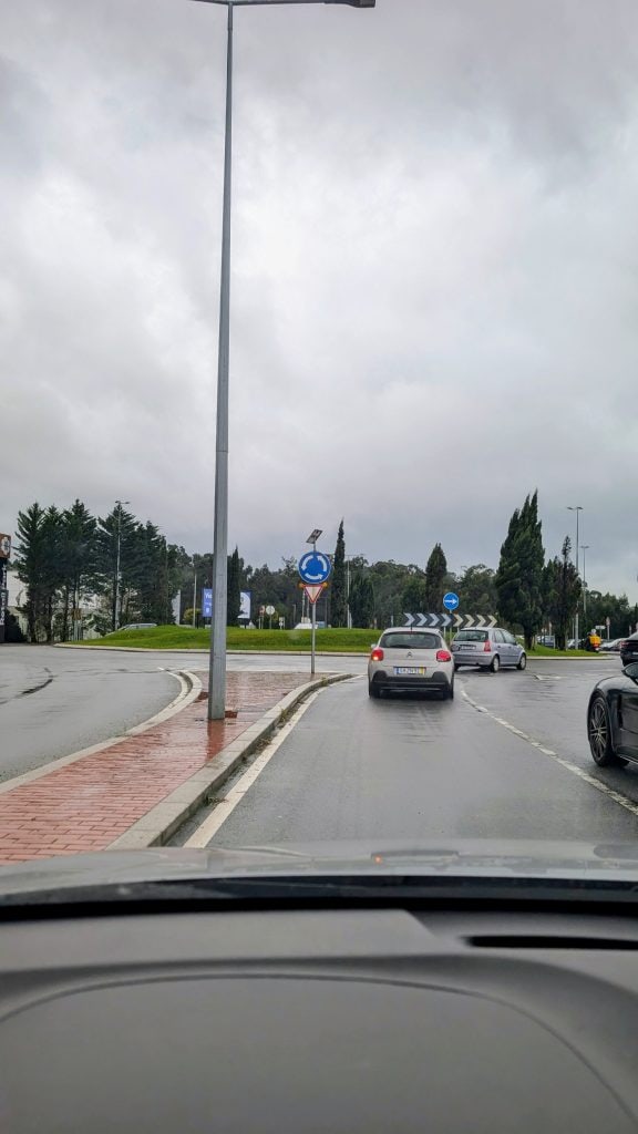 Cars approach a roundabout on a cloudy day, seen from inside another vehicle. The road is wet and bordered by a brick sidewalk and trees in the background.