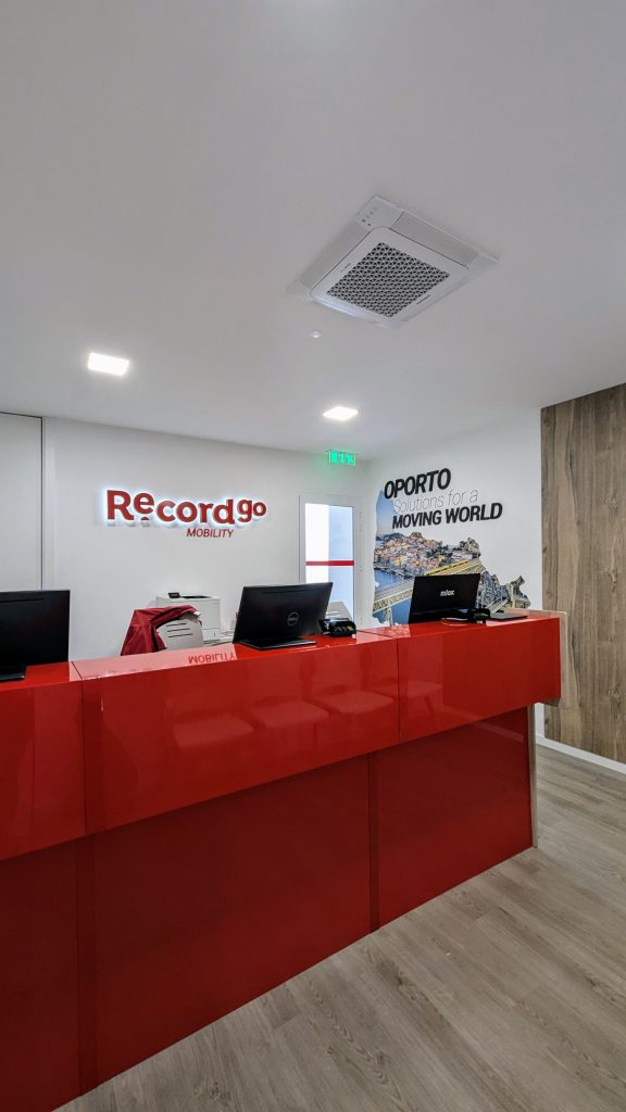 Red reception desk with two computers in a modern office. "Record go Mobility" and "Oporto Moving World" are visible on the white wall. Light wood flooring and wall decor shown.