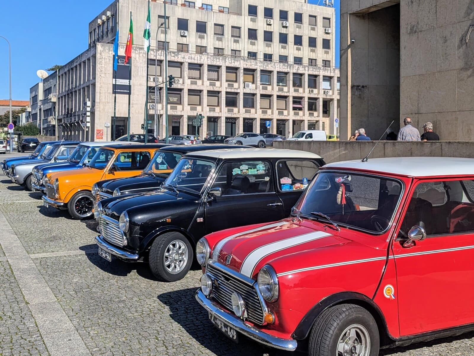 A row of classic Mini Coopers in various colors is parked on a cobblestone street in front of a modern office building.