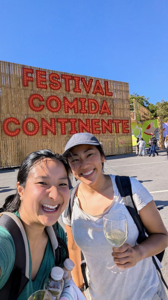 Two women smiling and posing with drinks in front of a large "Festival Comida Continente" sign at an outdoor event on a sunny day.