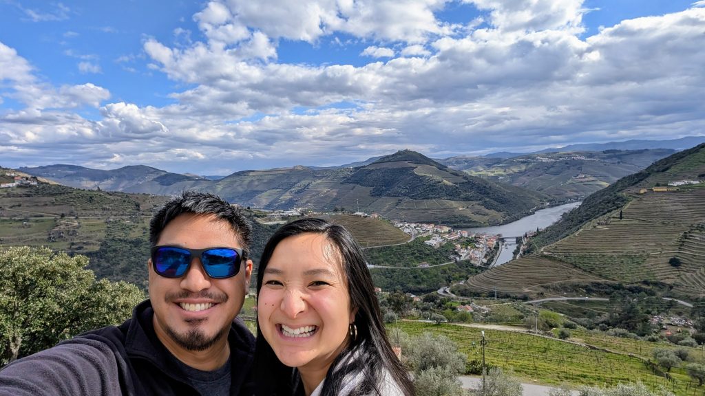 Two people smiling in the foreground with terraced green hills, a river, and a cloudy sky in the background.