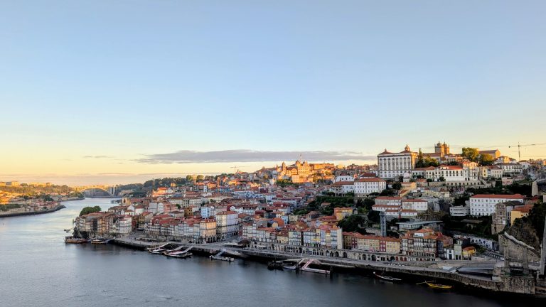 View of a city with colorful buildings along the riverbank under a clear sky, with a bridge visible in the background and boats docked by the water.