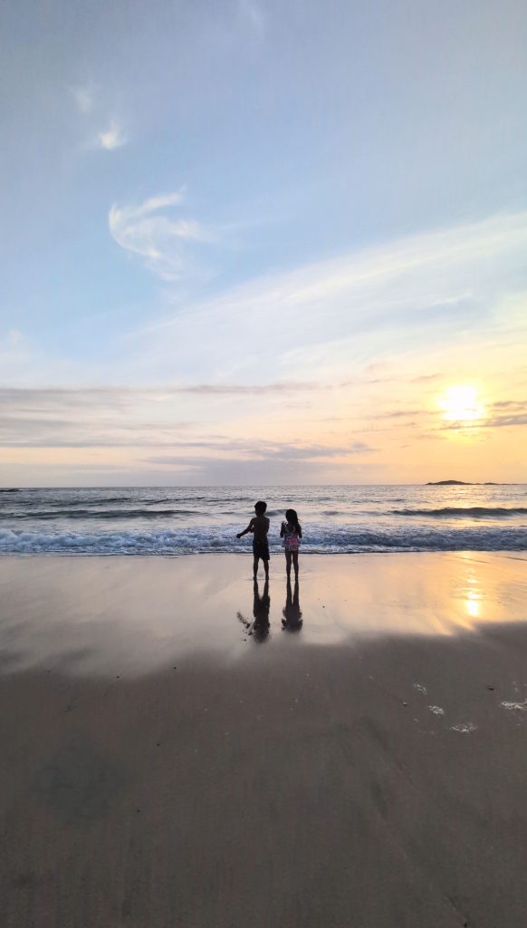 Two children stand on a sandy beach near the water's edge at sunset, facing the ocean with their reflections visible on the wet sand.