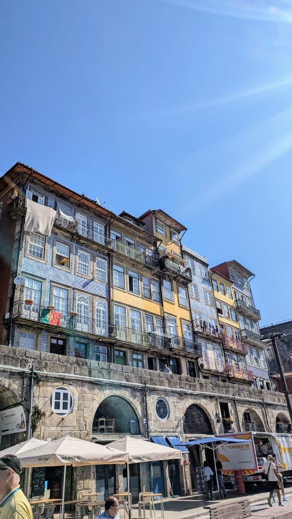 A row of colorful, old buildings with balconies and laundry hanging above street-level shops and cafes under a clear blue sky.