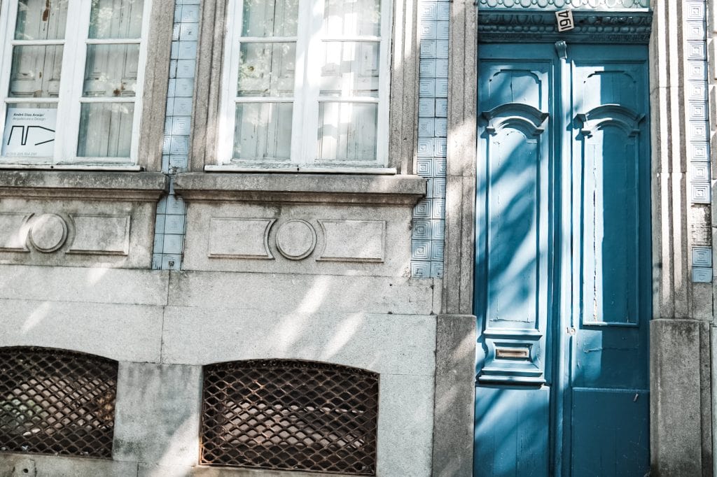Close-up of a building facade with two windows and a bright blue door; light and shadow pattern on the stone and blue tiles.