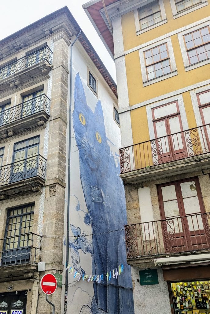 A large mural of a blue cat with yellow eyes is painted on the side of a building between two older, multi-story structures with balconies.