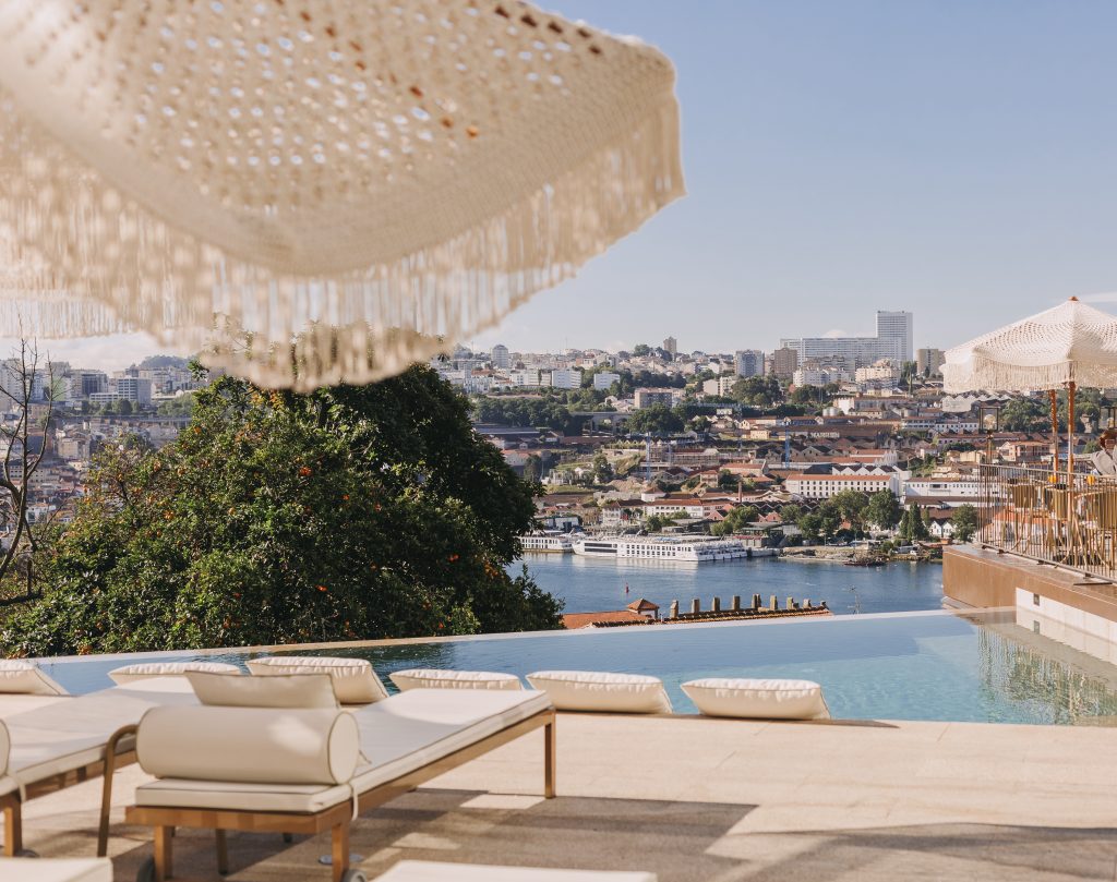 Lounge chairs by an infinity pool overlook a cityscape and river, with large fringed sun umbrellas providing shade under a clear sky.