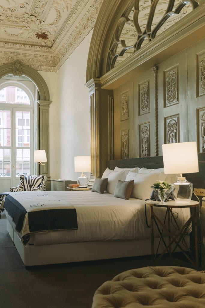 A hotel room with ornate ceilings, a large bed with white linens, two bedside tables with lamps, a zebra-patterned chair, and a large arched window letting in natural light.
