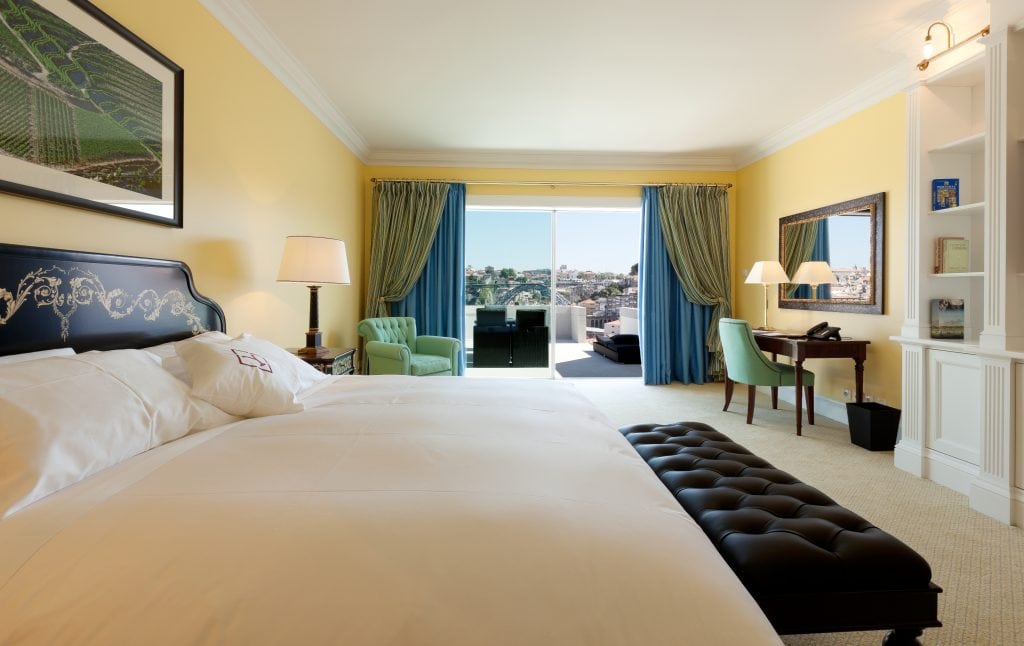 Hotel room with a large bed, black bench, armchairs, desk, bookshelves, and open balcony door revealing a city view under daylight.