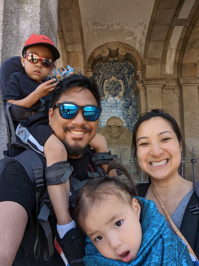A family of four poses for a selfie in front of a stone archway with tile artwork. A father carries a child on his shoulders, while a mother holds another child in a wrap carrier.
