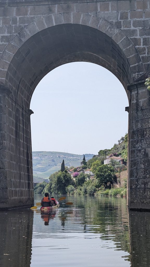 Two people in a kayak paddling on a river under a large stone bridge arch, with hills and houses in the distance.